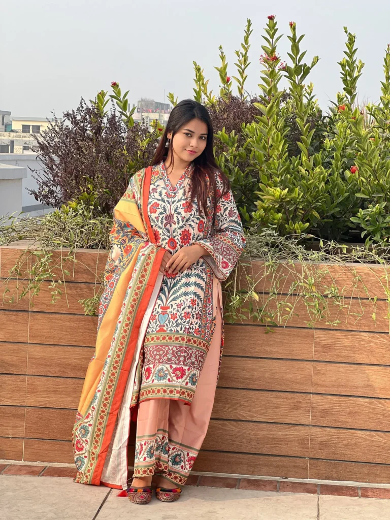 A woman is standing outdoors beside a wooden planter filled with lush green plants and small red flowers. She is wearing a traditional South Asian outfit in peach, orange, and red tones with intricate floral and geometric patterns. Her ensemble includes a long kurta, wide-legged trousers, and a matching dupatta draped over her shoulder. She has long, straight hair and is wearing colorful traditional footwear. The setting is bright and sunny with a clear sky in the background.