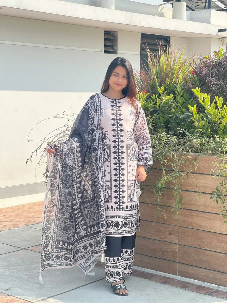 A woman is standing outdoors on a sunny day, next to a wooden planter filled with green plants and flowers. She is wearing a traditional South Asian outfit in black and white, featuring intricate geometric and floral patterns. Her ensemble includes a long kameez, matching trousers, and a coordinated printed dupatta. She has long, straight hair and is wearing black open-toe sandals. The background shows a modern building and a tiled floor.