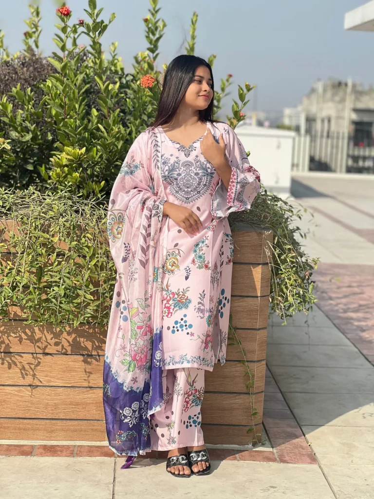 A woman is standing outdoors on a sunny day, in front of a wooden planter with green plants and red flowers. She is wearing a pastel pink traditional South Asian outfit adorned with floral and paisley prints in shades of blue, purple, and green. The outfit includes a long kurta, matching trousers, and a coordinating dupatta draped over her shoulder. She has long, straight hair and is wearing black sandals. In the background, a cityscape and blue sky are visible.