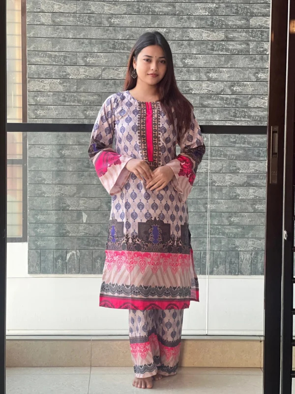 A woman is standing indoors in front of a modern gray brick wall, wearing a traditional South Asian outfit. The outfit is a printed kurta and matching trousers featuring a pattern of blue and purple motifs with accents of bright pink and black. The kurta has decorative borders and flared sleeves with pink detailing. She has long, straight hair, is barefoot, and wearing traditional jhumka earrings. She has a small bindi on her forehead and a calm expression on her face.