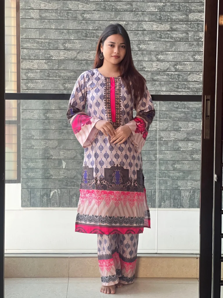 A woman is standing indoors in front of a modern gray brick wall, wearing a traditional South Asian outfit. The outfit is a printed kurta and matching trousers featuring a pattern of blue and purple motifs with accents of bright pink and black. The kurta has decorative borders and flared sleeves with pink detailing. She has long, straight hair, is barefoot, and wearing traditional jhumka earrings. She has a small bindi on her forehead and a calm expression on her face.