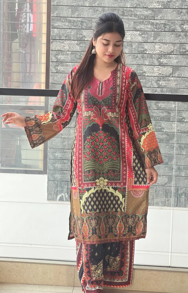 A woman is standing indoors in front of a textured gray brick wall, wearing a traditional South Asian outfit. The outfit is a vibrant printed kurta and matching trousers featuring intricate patterns in red, black, gold, and green with floral and geometric motifs. The kurta has long sleeves with detailed designs and a neckline with a keyhole cut. She has long, straight hair, partially tied back, and is wearing traditional jhumka earrings. Her expression is calm, and she is looking downward with one arm slightly extended.