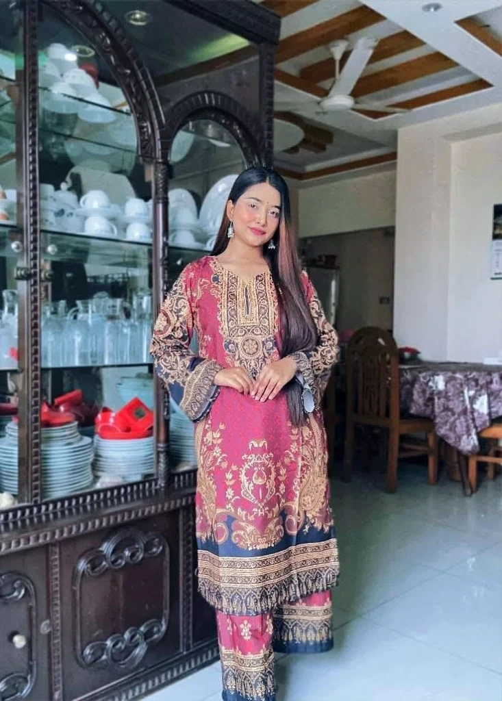 A woman is standing indoors near a dark wooden china cabinet filled with dishes and glassware. She is dressed in a maroon and navy blue traditional South Asian outfit featuring intricate golden patterns and ornate designs. The outfit includes a long kurta and matching straight pants. Her long straight hair is left open, and she is wearing dangling earrings. The room has white walls, a ceiling fan, and a dining area with wooden furniture in the background. The woman is smiling slightly and has a composed, elegant demeanor.