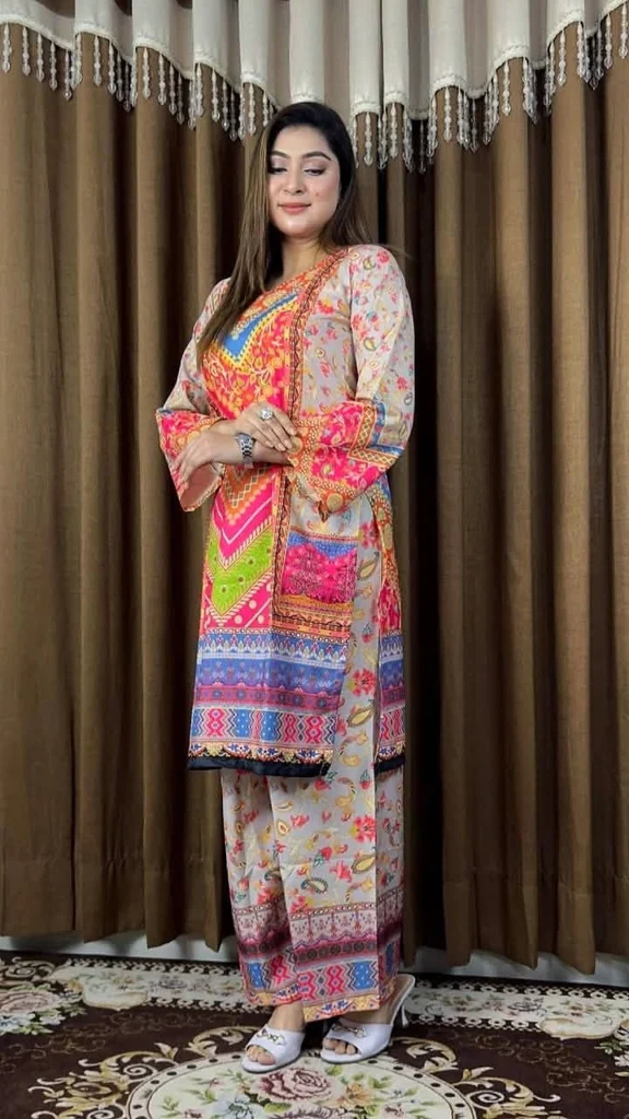 A woman is posing indoors in front of brown curtains with white and beige valances. She is wearing a vibrant traditional South Asian outfit featuring a multicolored kurta with intricate patterns in shades of pink, orange, green, blue, and yellow. The kurta has matching straight pants and a coordinating dupatta draped over one shoulder. Her outfit is adorned with floral and geometric prints. She is wearing white heeled sandals, rings, and a wristwatch, and her straight hair is left open. The room has a patterned rug beneath her feet, adding to the warm, homely ambiance.