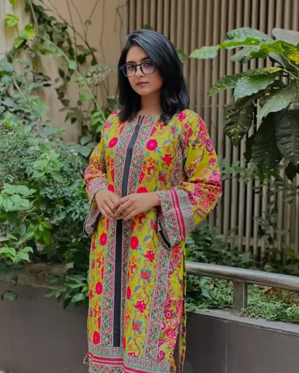 A woman stands outdoors near a garden area, surrounded by lush green plants and a beige wall with vertical slats. She is wearing a bright yellow traditional kurta adorned with vibrant floral prints in red, pink, blue, and green. The kurta features vertical embroidered panels with intricate pink and green patterns along the front and sleeves. She has shoulder-length black hair and is wearing eyeglasses. Her hands are gently clasped in front of her, and her expression is calm and poised.