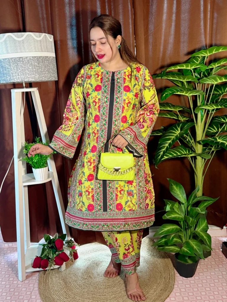 A woman stands indoors on a round jute rug, dressed in a beige traditional outfit with navy blue, pink, and black patterns. She holds a novelty clutch shaped like bundles of currency notes. Her long, straight hair is left open, and she wears bright pink lipstick. The setting includes brown curtains, a white tripod lamp with a potted plant, a bunch of red roses on the floor, and a tall artificial green plant on the right.