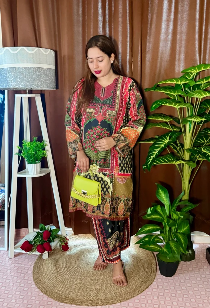 A woman stands barefoot on a round jute rug, wearing a vibrant traditional outfit featuring a mix of red, green, black, and gold ethnic patterns. She carries a bright lime green handbag with a gold chain strap. Her long, straight hair is worn down, and she has red lipstick. The background includes brown curtains, a white tripod lamp with a potted plant on a shelf, a cluster of red roses on the floor, and a large leafy artificial plant on the right.
