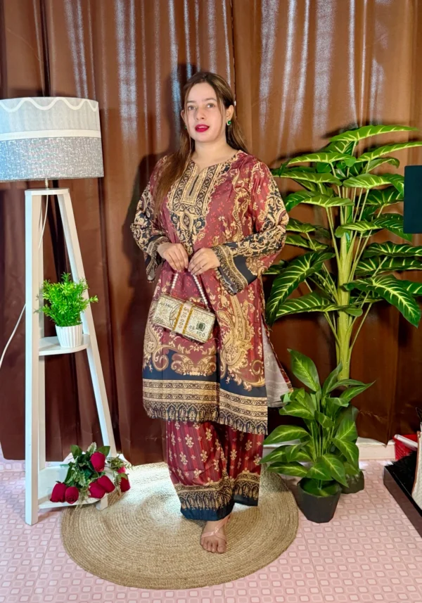 A woman stands indoors on a round jute rug, dressed in a beige traditional outfit with navy blue, pink, and black patterns. She holds a novelty clutch shaped like bundles of currency notes. Her long, straight hair is left open, and she wears bright pink lipstick. The setting includes brown curtains, a white tripod lamp with a potted plant, a bunch of red roses on the floor, and a tall artificial green plant on the right.