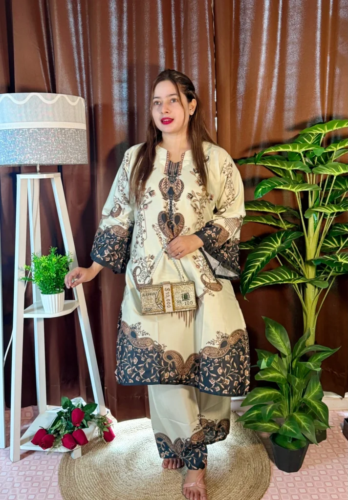 A woman is standing indoors on a circular jute rug, dressed in a cream and dark brown printed traditional kurti and matching pants with peacock and floral motifs. She is holding a gold beaded clutch shaped like a bundle of currency notes. Her hair is straight and parted to the side, and she is barefoot. The background includes brown curtains, a white tripod lamp with a potted plant on a shelf, a green artificial plant, and a few red roses placed on the floor.