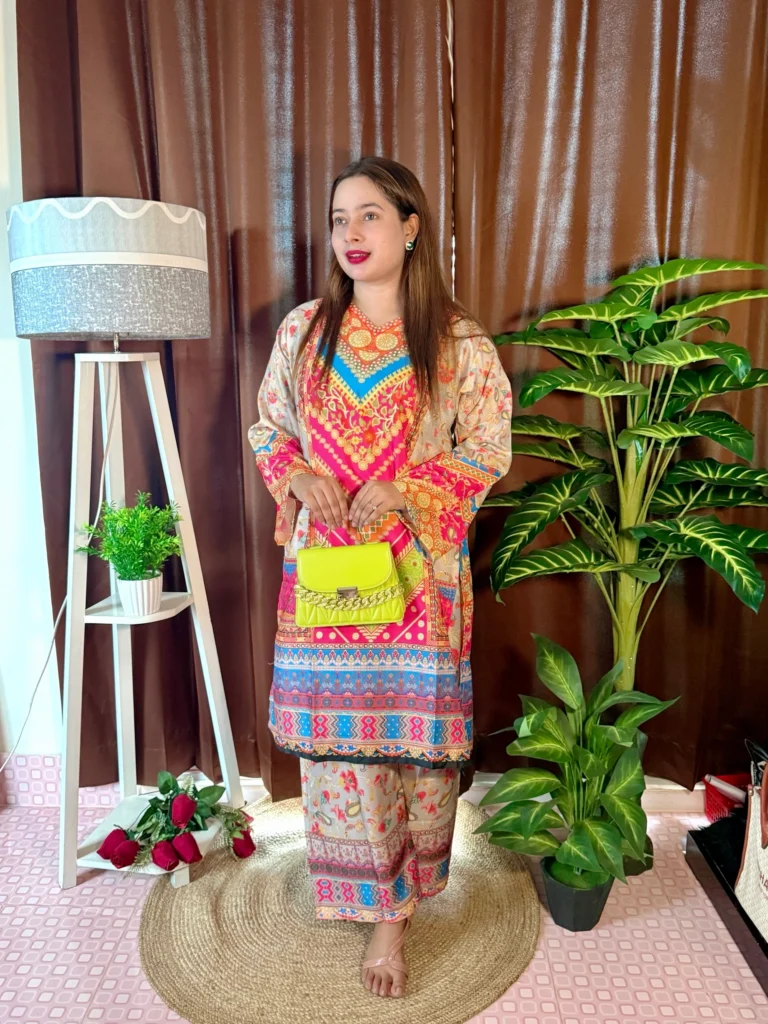 A woman stands indoors on a round jute rug, dressed in a beige traditional outfit with navy blue, pink, and black patterns. She holds a novelty clutch shaped like bundles of currency notes. Her long, straight hair is left open, and she wears bright pink lipstick. The setting includes brown curtains, a white tripod lamp with a potted plant, a bunch of red roses on the floor, and a tall artificial green plant on the right.