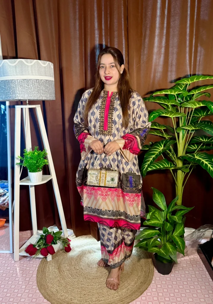 A woman poses barefoot on a round jute rug, wearing a traditional cream and navy printed kurti and matching trousers with pink and black accents. She carries a novelty clutch styled like a stack of dollar bills. Her hair is long and worn down, and she has bright pink lipstick. The background features brown curtains, a white tripod lamp with a potted plant on its shelf, a bunch of red roses on the floor, and a large artificial green plant on the right.