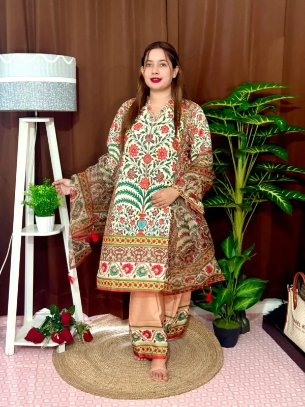Woman dressed in a vibrant traditional South Asian outfit featuring a mix of peach, orange, and red tones with intricate floral and geometric with ornate patterns and floral motifs. She holds a sheer printed dupatta with maroon and green accents. She stands barefoot on a circular jute rug, with indoor plants and a white lampstand beside her, against a brown curtain backdrop. A few red roses are placed on the floor near the lampstand.