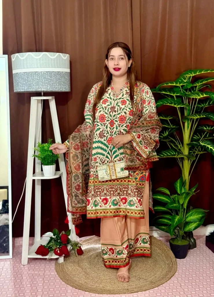 Woman dressed in a vibrant traditional South Asian outfit featuring a mix of peach, orange, and red tones with intricate floral and geometric with ornate patterns and floral motifs. She holds a sheer printed dupatta with maroon and green accents. She stands barefoot on a circular jute rug, with indoor plants and a white lampstand beside her, against a brown curtain backdrop. A few red roses are placed on the floor near the lampstand.