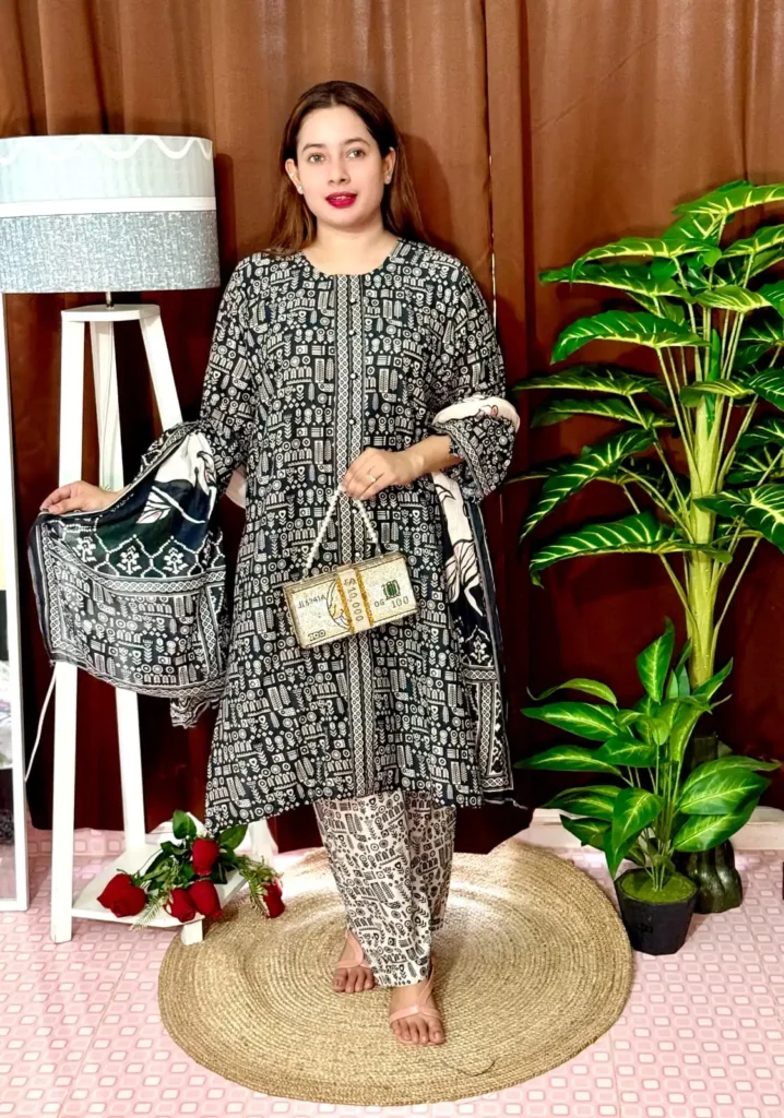 Woman wearing a black and white traditional South Asian outfit with intricate geometric and tribal-style patterns. She holds a matching printed dupatta in one hand and carries a novelty handbag designed like a bundle of US $100 bills. She stands barefoot on a circular jute rug, with indoor plants and a white lampstand beside her, against a brown curtain backdrop. A few red roses are placed on the floor near the lampstand.