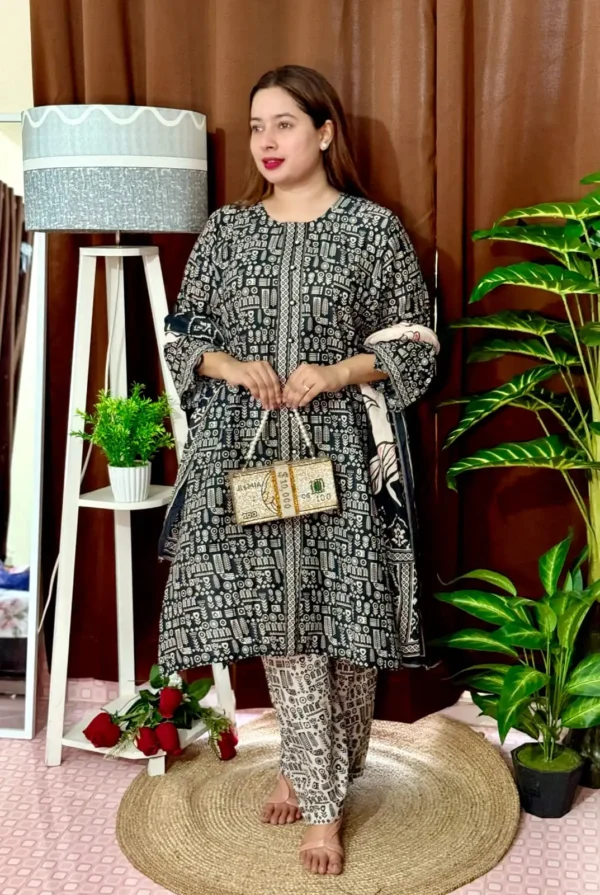 Woman wearing a black and white traditional South Asian outfit with intricate geometric and tribal-style patterns. She holds a matching printed dupatta in one hand and carries a novelty handbag designed like a bundle of US $100 bills. She stands barefoot on a circular jute rug, with indoor plants and a white lampstand beside her, against a brown curtain backdrop. A few red roses are placed on the floor near the lampstand.