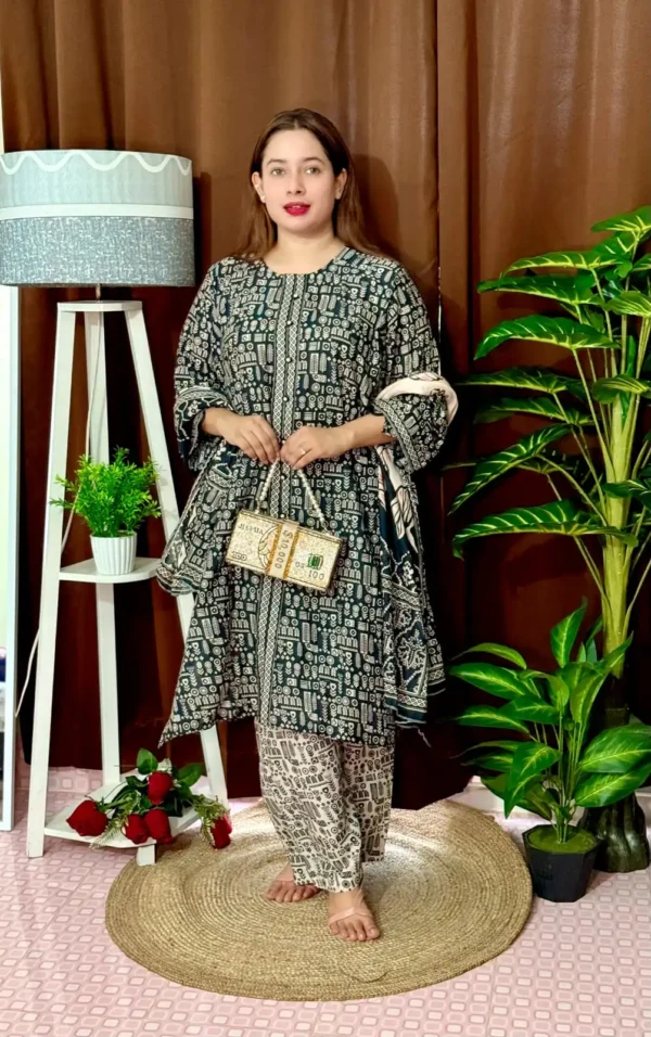Woman wearing a black and white traditional South Asian outfit with intricate geometric and tribal-style patterns. She holds a matching printed dupatta in one hand and carries a novelty handbag designed like a bundle of US $100 bills. She stands barefoot on a circular jute rug, with indoor plants and a white lampstand beside her, against a brown curtain backdrop. A few red roses are placed on the floor near the lampstand.