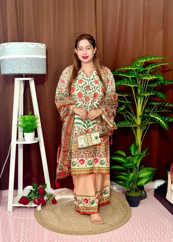 Woman dressed in a vibrant traditional South Asian outfit featuring a mix of peach, orange, and red tones with intricate floral and geometric with ornate patterns and floral motifs. She holds a sheer printed dupatta with maroon and green accents. She stands barefoot on a circular jute rug, with indoor plants and a white lampstand beside her, against a brown curtain backdrop. A few red roses are placed on the floor near the lampstand.