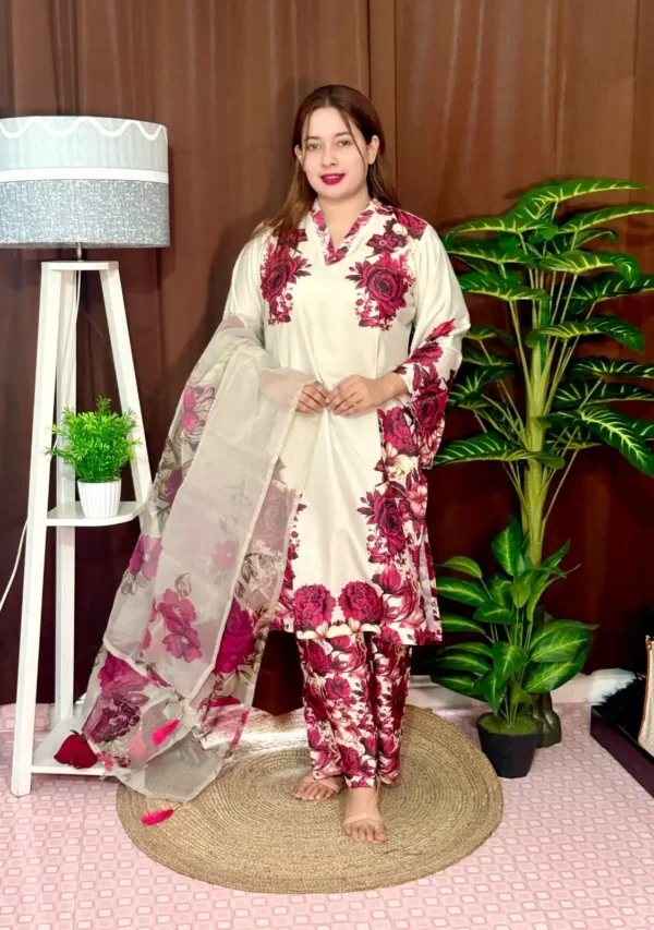 Woman dressed in a vibrant traditional South Asian outfit featuring a mix of pink and white with ornate patterns and floral motifs. She holds a sheer printed dupatta with maroon and green accents. She stands barefoot on a circular jute rug, with indoor plants and a white lampstand beside her, against a brown curtain backdrop. A few red roses are placed on the floor near the lampstand.