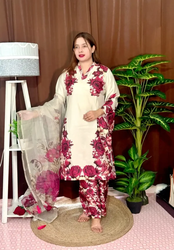 Woman dressed in a vibrant traditional South Asian outfit featuring a mix of pink and white with ornate patterns and floral motifs. She holds a sheer printed dupatta with maroon and green accents. She stands barefoot on a circular jute rug, with indoor plants and a white lampstand beside her, against a brown curtain backdrop. A few red roses are placed on the floor near the lampstand.