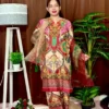 Woman dressed in a vibrant traditional South Asian outfit featuring a mix of red, pink, green, gold, and cream with ornate patterns and floral motifs. She holds a sheer printed dupatta with maroon and green accents. She stands barefoot on a circular jute rug, with indoor plants and a white lampstand beside her, against a brown curtain backdrop. A few red roses are placed on the floor near the lampstand.