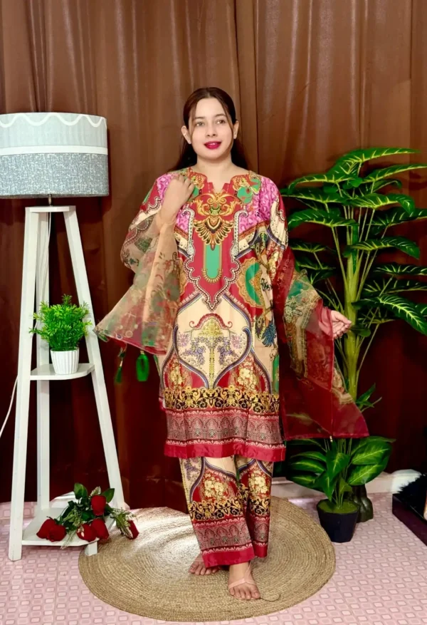 Woman dressed in a vibrant traditional South Asian outfit featuring a mix of red, pink, green, gold, and cream with ornate patterns and floral motifs. She holds a sheer printed dupatta with maroon and green accents. She stands barefoot on a circular jute rug, with indoor plants and a white lampstand beside her, against a brown curtain backdrop. A few red roses are placed on the floor near the lampstand.