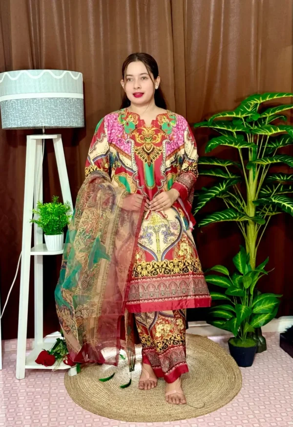 Woman dressed in a vibrant traditional South Asian outfit featuring a mix of red, pink, green, gold, and cream with ornate patterns and floral motifs. She holds a sheer printed dupatta with maroon and green accents. She stands barefoot on a circular jute rug, with indoor plants and a white lampstand beside her, against a brown curtain backdrop. A few red roses are placed on the floor near the lampstand.