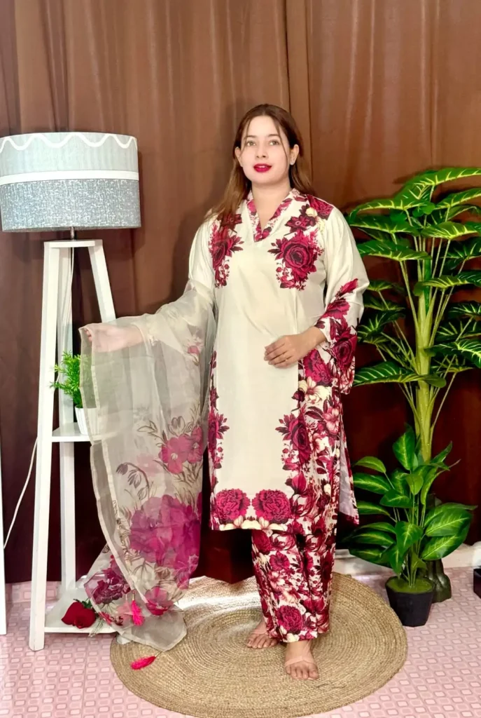 Woman dressed in a vibrant traditional South Asian outfit featuring a mix of pink and white with ornate patterns and floral motifs. She holds a sheer printed dupatta with maroon and green accents. She stands barefoot on a circular jute rug, with indoor plants and a white lampstand beside her, against a brown curtain backdrop. A few red roses are placed on the floor near the lampstand.