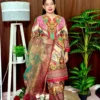 Woman dressed in a vibrant traditional South Asian outfit featuring a mix of red, pink, green, gold, and cream with ornate patterns and floral motifs. She holds a sheer printed dupatta with maroon and green accents. She stands barefoot on a circular jute rug, with indoor plants and a white lampstand beside her, against a brown curtain backdrop. A few red roses are placed on the floor near the lampstand.