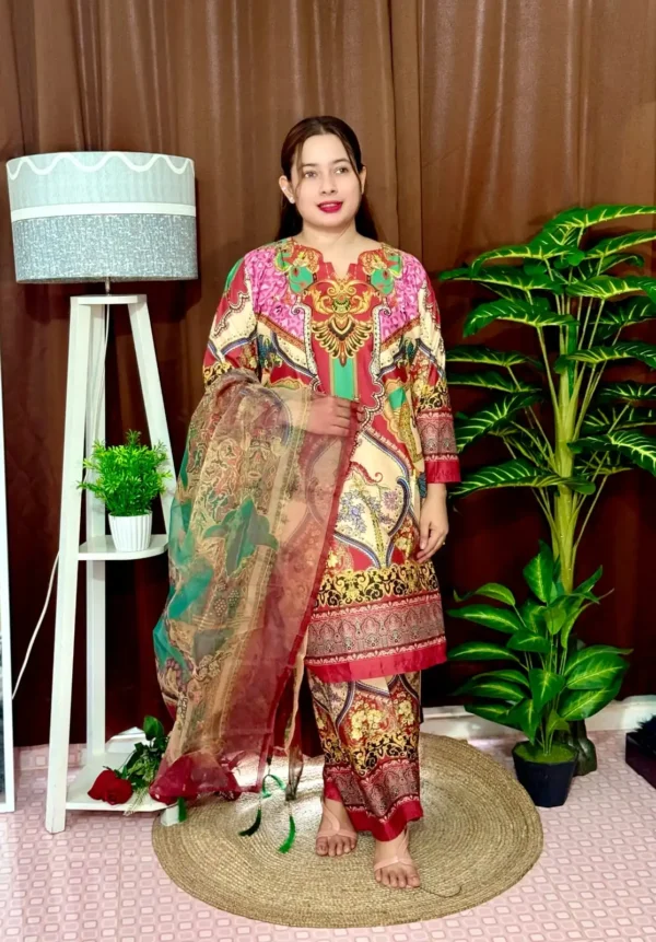 Woman dressed in a vibrant traditional South Asian outfit featuring a mix of red, pink, green, gold, and cream with ornate patterns and floral motifs. She holds a sheer printed dupatta with maroon and green accents. She stands barefoot on a circular jute rug, with indoor plants and a white lampstand beside her, against a brown curtain backdrop. A few red roses are placed on the floor near the lampstand.