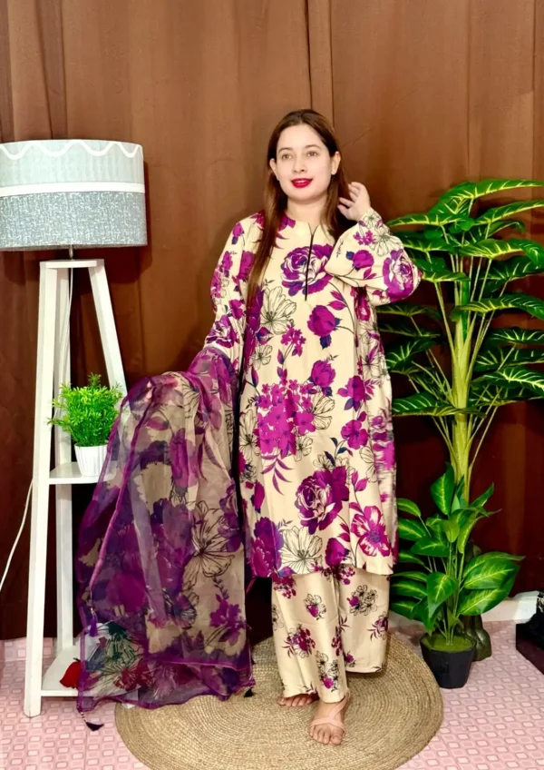 A woman stands indoors wearing a beige traditional South Asian outfit with vibrant purple and black floral prints. She holds a matching chiffon dupatta with similar floral patterns. The outfit consists of a long kameez with full sleeves and loose trousers. She has long straight brown hair and wears pink lipstick. The background features a brown curtain, a tall white lamp with a grey shade, a white shelf with a potted plant, and large green artificial plants.