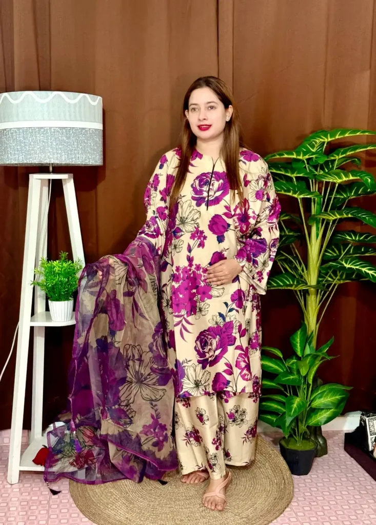 A woman stands indoors wearing a beige traditional South Asian outfit with vibrant purple and black floral prints. She holds a matching chiffon dupatta with similar floral patterns. The outfit consists of a long kameez with full sleeves and loose trousers. She has long straight brown hair and wears pink lipstick. The background features a brown curtain, a tall white lamp with a grey shade, a white shelf with a potted plant, and large green artificial plants.