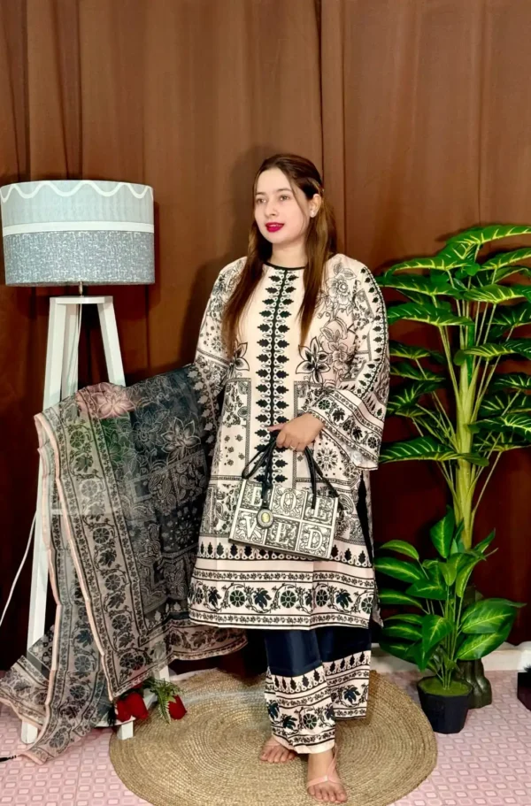 A woman stands indoors wearing a traditional South Asian outfit in beige and black with intricate floral and geometric patterns. The outfit includes a long kameez with printed detailing, straight trousers with matching prints, and a printed dupatta draped over her arm. She holds a black and white handbag that reads "LOVED." Her hair is straight and loose, and she wears bright pink lipstick. The background includes a brown curtain, a white standing lamp, a small potted plant on a white shelf, and tall green artificial plants.