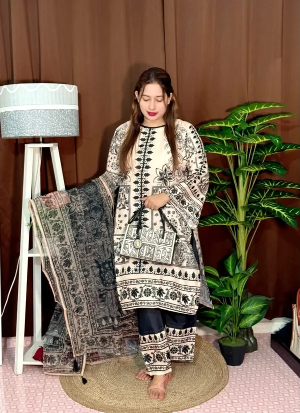 A woman stands indoors wearing a traditional South Asian outfit in beige and black with intricate floral and geometric patterns. The outfit includes a long kameez with printed detailing, straight trousers with matching prints, and a printed dupatta draped over her arm. She holds a black and white handbag that reads "LOVED." Her hair is straight and loose, and she wears bright pink lipstick. The background includes a brown curtain, a white standing lamp, a small potted plant on a white shelf, and tall green artificial plants.
