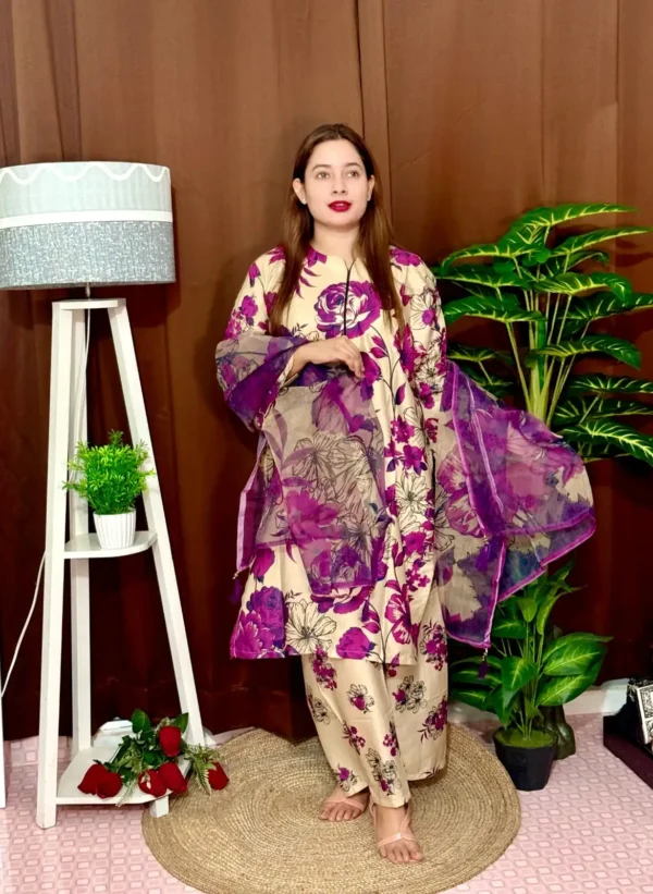 A woman stands indoors wearing a beige traditional South Asian outfit with vibrant purple and black floral prints. She holds a matching chiffon dupatta with similar floral patterns. The outfit consists of a long kameez with full sleeves and loose trousers. She has long straight brown hair and wears pink lipstick. The background features a brown curtain, a tall white lamp with a grey shade, a white shelf with a potted plant, and large green artificial plants.