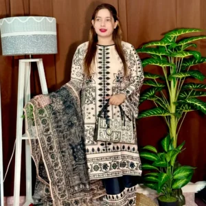 A woman stands indoors wearing a traditional South Asian outfit in beige and black with intricate floral and geometric patterns. The outfit includes a long kameez with printed detailing, straight trousers with matching prints, and a printed dupatta draped over her arm. She holds a black and white handbag that reads "LOVED." Her hair is straight and loose, and she wears bright pink lipstick. The background includes a brown curtain, a white standing lamp, a small potted plant on a white shelf, and tall green artificial plants.