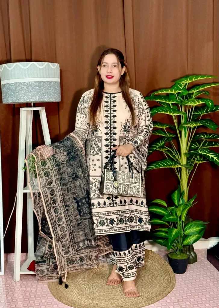 A woman stands indoors wearing a traditional South Asian outfit in beige and black with intricate floral and geometric patterns. The outfit includes a long kameez with printed detailing, straight trousers with matching prints, and a printed dupatta draped over her arm. She holds a black and white handbag that reads "LOVED." Her hair is straight and loose, and she wears bright pink lipstick. The background includes a brown curtain, a white standing lamp, a small potted plant on a white shelf, and tall green artificial plants.