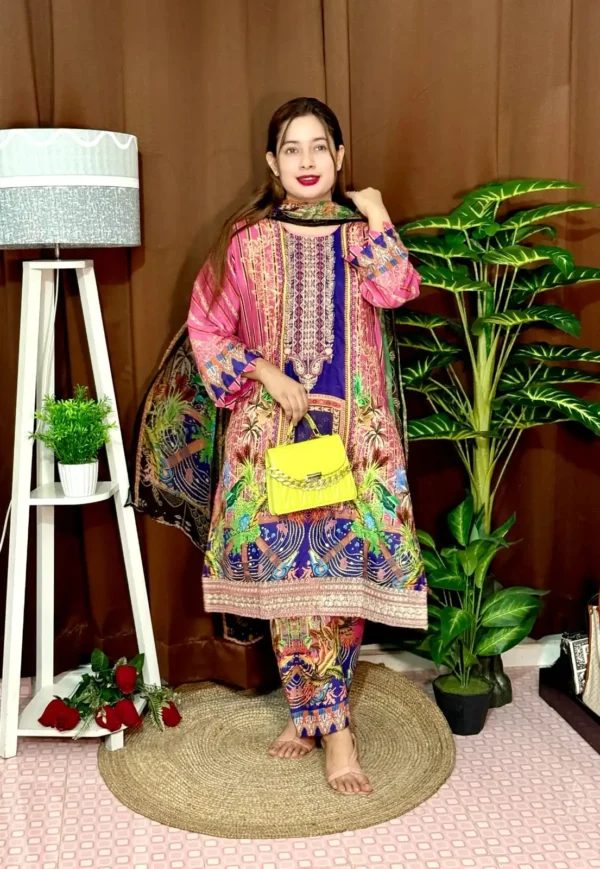 Woman wearing a colorful traditional South Asian outfit featuring a vibrant mix of pink, blue, and green with intricate patterns. She holds part of her printed dupatta in one hand and carries a small yellow handbag with a chain strap. She stands barefoot on a round jute rug, with indoor plants and a white lampstand beside her, against a brown curtain backdrop. A few red roses are placed on the floor near the lampstand.