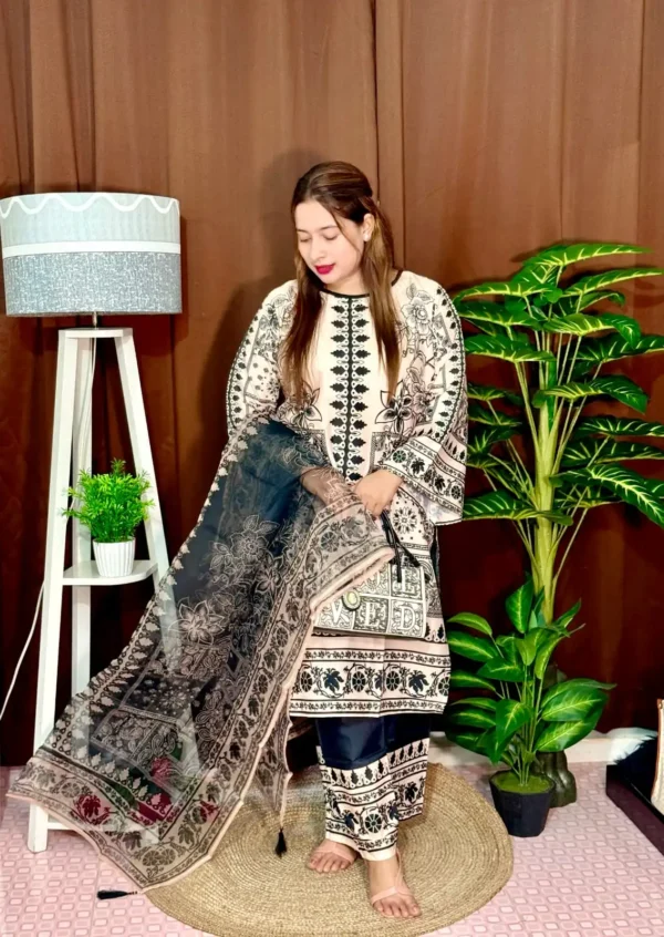 A woman stands indoors wearing a traditional South Asian outfit in beige and black with intricate floral and geometric patterns. The outfit includes a long kameez with printed detailing, straight trousers with matching prints, and a printed dupatta draped over her arm. She holds a black and white handbag that reads "LOVED." Her hair is straight and loose, and she wears bright pink lipstick. The background includes a brown curtain, a white standing lamp, a small potted plant on a white shelf, and tall green artificial plants.