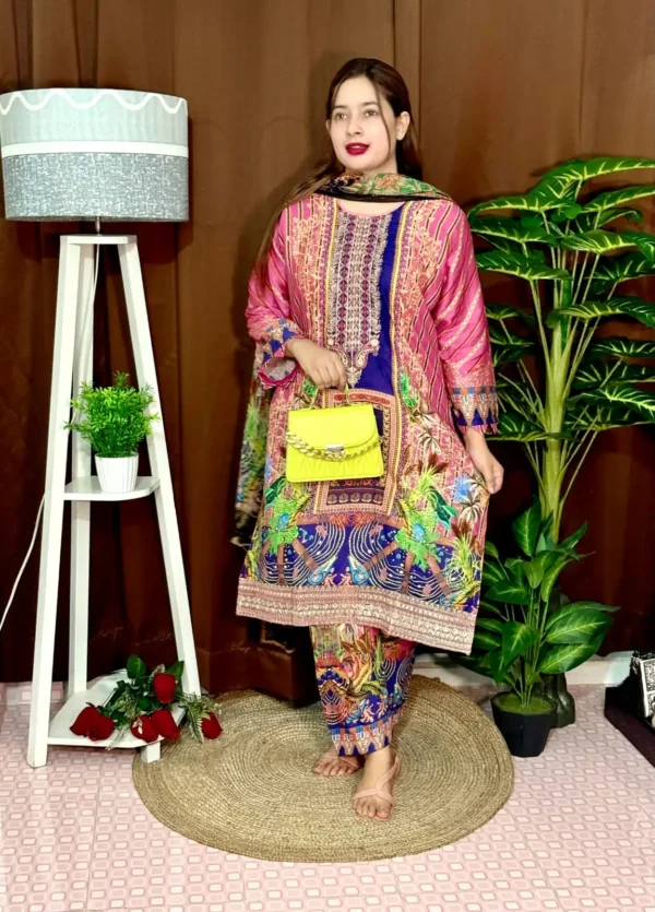 Woman wearing a colorful traditional South Asian outfit featuring a vibrant mix of pink, blue, and green with intricate patterns. She holds part of her printed dupatta in one hand and carries a small yellow handbag with a chain strap. She stands barefoot on a round jute rug, with indoor plants and a white lampstand beside her, against a brown curtain backdrop. A few red roses are placed on the floor near the lampstand.