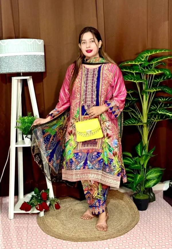 Woman wearing a colorful traditional South Asian outfit featuring a vibrant mix of pink, blue, and green with intricate patterns. She holds part of her printed dupatta in one hand and carries a small yellow handbag with a chain strap. She stands barefoot on a round jute rug, with indoor plants and a white lampstand beside her, against a brown curtain backdrop. A few red roses are placed on the floor near the lampstand.