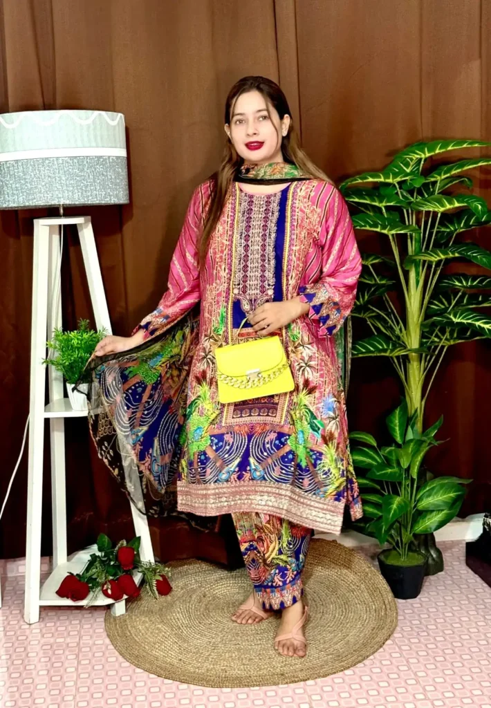 Woman wearing a colorful traditional South Asian outfit featuring a vibrant mix of pink, blue, and green with intricate patterns. She holds part of her printed dupatta in one hand and carries a small yellow handbag with a chain strap. She stands barefoot on a round jute rug, with indoor plants and a white lampstand beside her, against a brown curtain backdrop. A few red roses are placed on the floor near the lampstand.