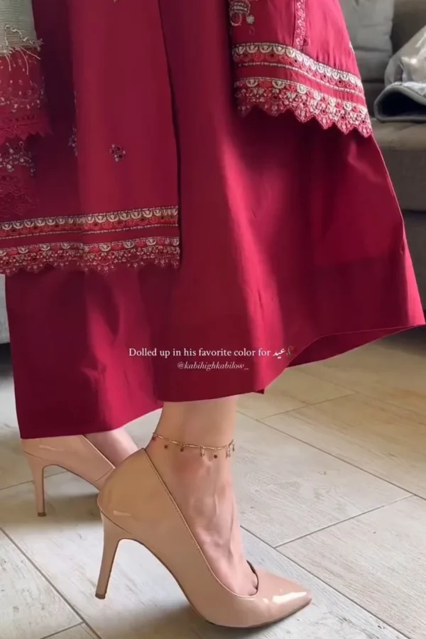 Woman wearing a deep red traditional embroidered kameez with floral and vine motifs in silver and light gold threads. The neckline features a scalloped lace design, and the long sleeves have intricate embroidery near the cuffs. She holds her phone in one hand, revealing a gold ring and a watch on the other wrist. Text on the image reads: "Dolled up in his favorite color for Eid." Let me know if you'd like a combined caption or product-style description for all three images.