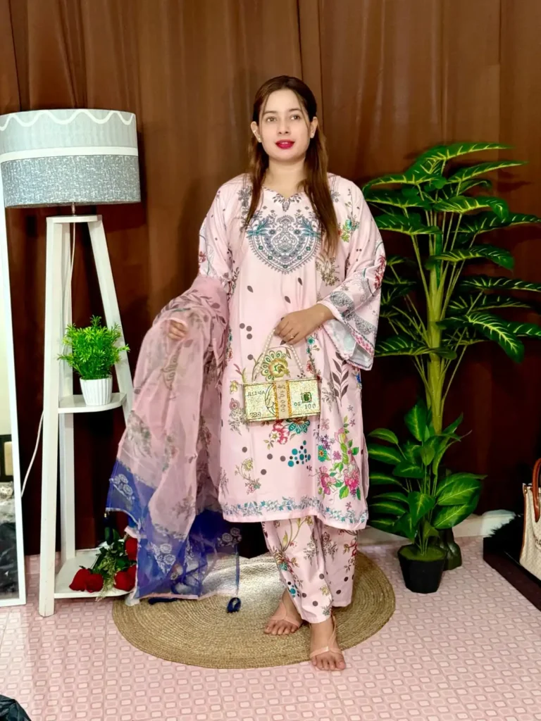 Woman dressed in a vibrant traditional South Asian outfit featuring a mix of pink and blue with ornate patterns and floral motifs. She holds a sheer printed dupatta with maroon and green accents. She stands barefoot on a circular jute rug, with indoor plants and a white lampstand beside her, against a brown curtain backdrop. A few red roses are placed on the floor near the lampstand.