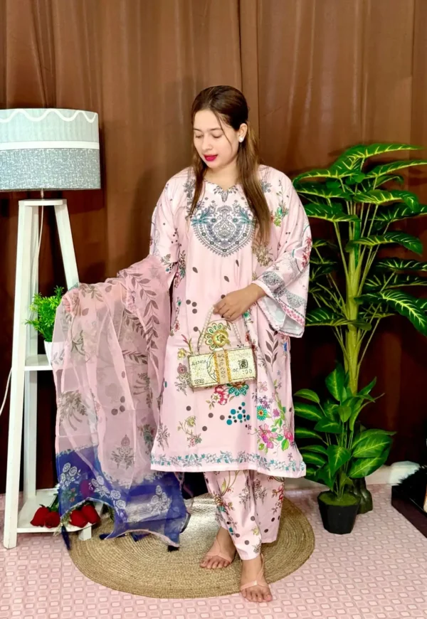 Woman dressed in a vibrant traditional South Asian outfit featuring a mix of pink and blue with ornate patterns and floral motifs. She holds a sheer printed dupatta with maroon and green accents. She stands barefoot on a circular jute rug, with indoor plants and a white lampstand beside her, against a brown curtain backdrop. A few red roses are placed on the floor near the lampstand.