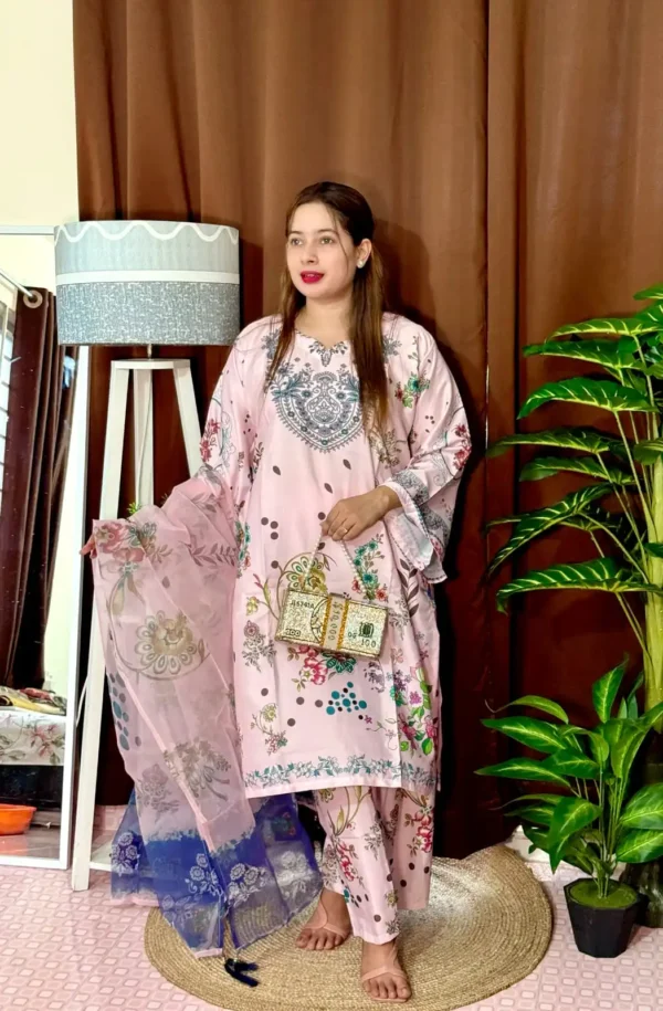 Woman dressed in a vibrant traditional South Asian outfit featuring a mix of pink and blue with ornate patterns and floral motifs. She holds a sheer printed dupatta with maroon and green accents. She stands barefoot on a circular jute rug, with indoor plants and a white lampstand beside her, against a brown curtain backdrop. A few red roses are placed on the floor near the lampstand.