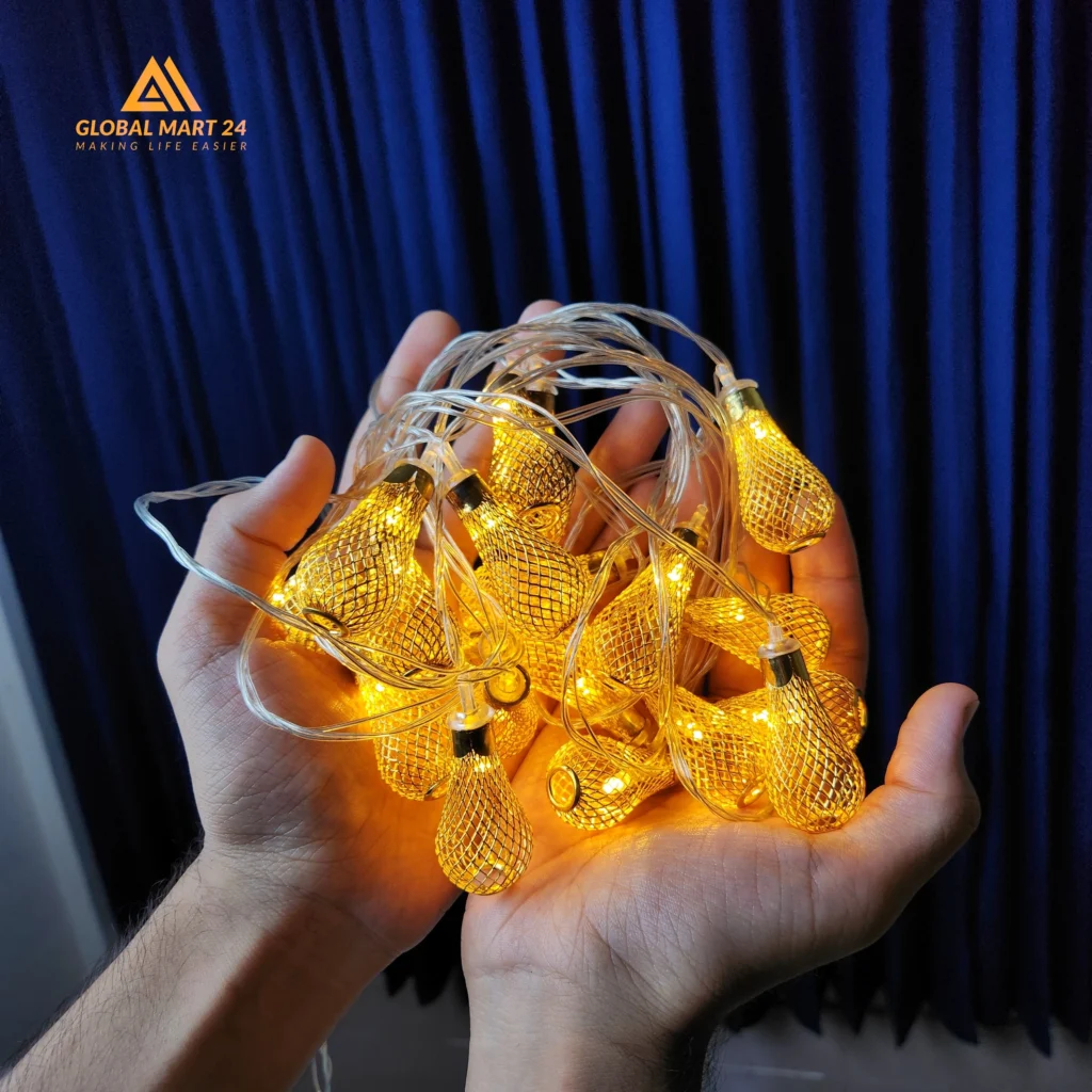 A pair of hands is holding a glowing string of decorative golden LED lights with cylindrical metal covers featuring a mesh design. The lights emit a warm yellow glow, and the background is a deep blue curtain. The logo of "GLOBAL MART 24" is visible in the upper left corner with the tagline "MAKING LIFE EASIER."