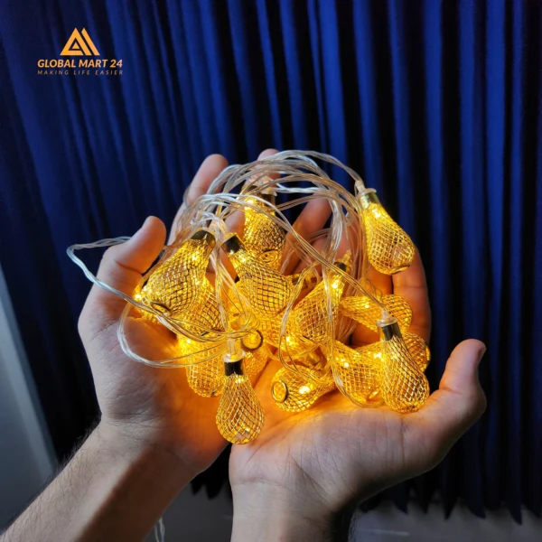 A pair of hands is holding a glowing string of decorative golden LED lights with cylindrical metal covers featuring a mesh design. The lights emit a warm yellow glow, and the background is a deep blue curtain. The logo of "GLOBAL MART 24" is visible in the upper left corner with the tagline "MAKING LIFE EASIER."
