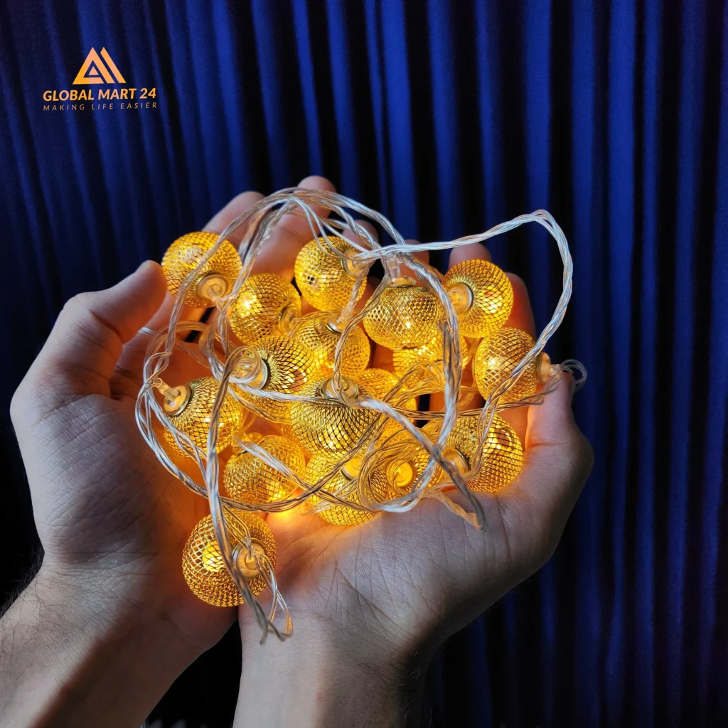 A pair of hands is holding a glowing string of decorative golden LED lights with cylindrical metal covers featuring a mesh design. The lights emit a warm yellow glow, and the background is a deep blue curtain. The logo of "GLOBAL MART 24" is visible in the upper left corner with the tagline "MAKING LIFE EASIER."