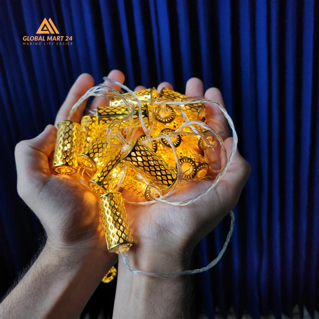 A pair of hands is holding a glowing string of decorative golden LED lights with cylindrical metal covers featuring a mesh design. The lights emit a warm yellow glow, and the background is a deep blue curtain. The logo of "GLOBAL MART 24" is visible in the upper left corner with the tagline "MAKING LIFE EASIER."