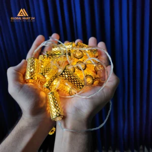 A pair of hands is holding a glowing string of decorative golden LED lights with cylindrical metal covers featuring a mesh design. The lights emit a warm yellow glow, and the background is a deep blue curtain. The logo of "GLOBAL MART 24" is visible in the upper left corner with the tagline "MAKING LIFE EASIER."